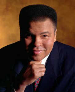 headshot of Muhammad Ali smiling in a suite and tie with right hand under chin