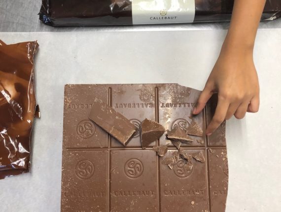 Big bar of chocolate with chunks broken off it and a hand reaching for one chunk