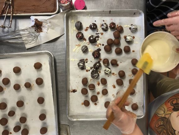 Chocolate truffles on a tray, while someone drizzles topping on them using a spatula