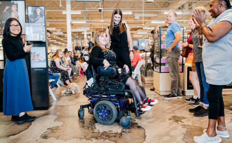 A PYD participant models in her wheelchair, with a PYD staff member behind her, smiling.